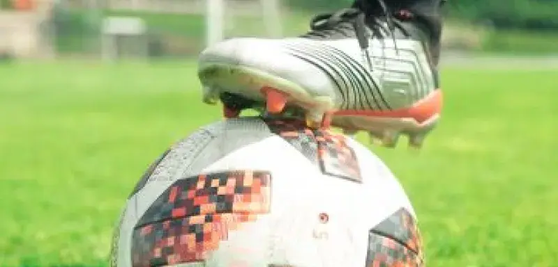 Close-up of a soccer ball with a player's foot on it, set on a vibrant green soccer field during a sunny day.