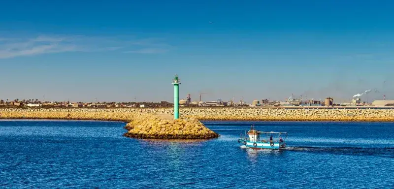 A small fishing boat sailing past a tall lighthouse in Gabès harbor, Tunisie.