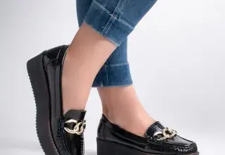 Gros plan sur des mocassins noirs à chaîne dorée portés avec un jean sur fond blanc