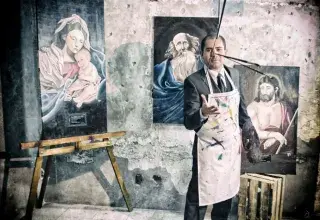 Artist in a suit and apron poses with Renaissance-style paintings in an old studio.