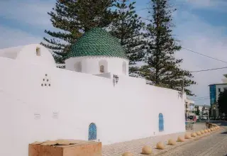 A whitewashed Mediterranean building with a green dome, surrounded by trees, and under a clear blue sky.