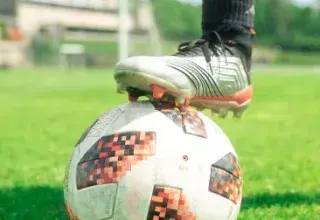 Close-up of a soccer ball with a player's foot on it, set on a vibrant green soccer field during a sunny day.