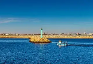 A small fishing boat sailing past a tall lighthouse in Gabès harbor, Tunisie.
