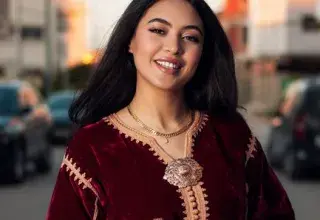 Elegant woman in a red velvet dress smiling on a street during sunset.